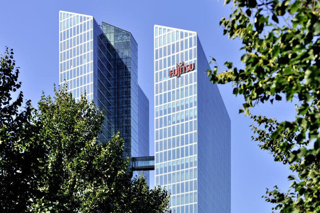 Zwei moderne Hochhaustürme mit Fujitsu-Logo an der Fassade, umgeben von Bäumen, vor blauem Himmel.