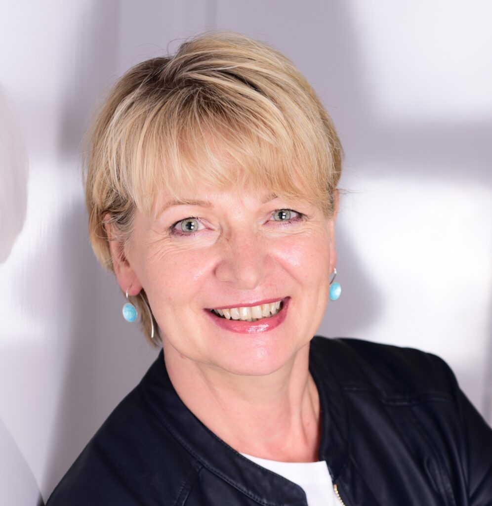Birgit Schreiber mit blondem, kurzem Haar und blauen Ohrringen vor hellem Hintergrund lächelt in die Kamera.