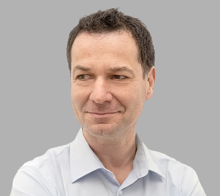Porträt von Andreas Roßmann mit kurzen dunklen Haaren in einem hellem Hemd, der leicht zur Seite blickt und dezent lächelt, vor grauem Hintergrund.