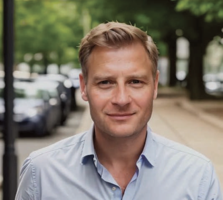 Frank Ponndorf in hellblauem Hemd steht auf einem städtischen Gehweg und blickt direkt in die Kamera, Bäume und Autos im Hintergrund.