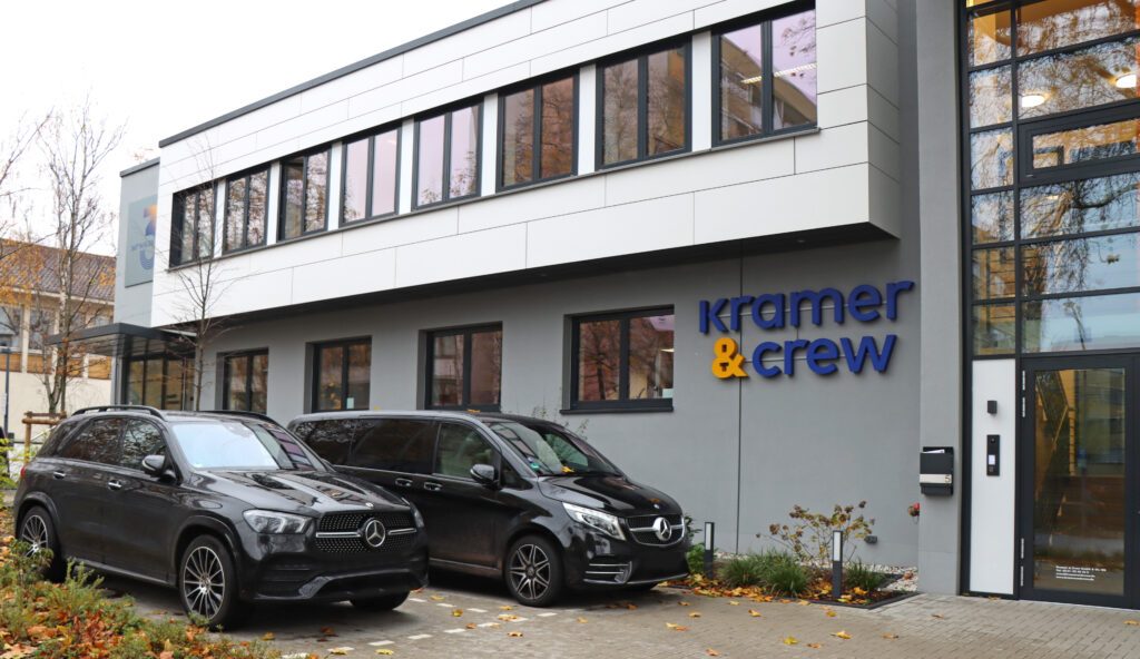 Bürogebäude von Kramer & Crew mit zwei schwarzen Firmenfahrzeugen davor, herbstliche Umgebung.