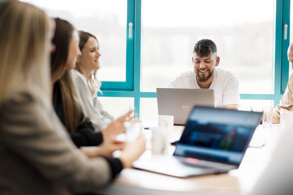 Teammeeting in einem hellen Büro mit mehreren Personen, im Fokus ein lachender Mann mit Laptop.