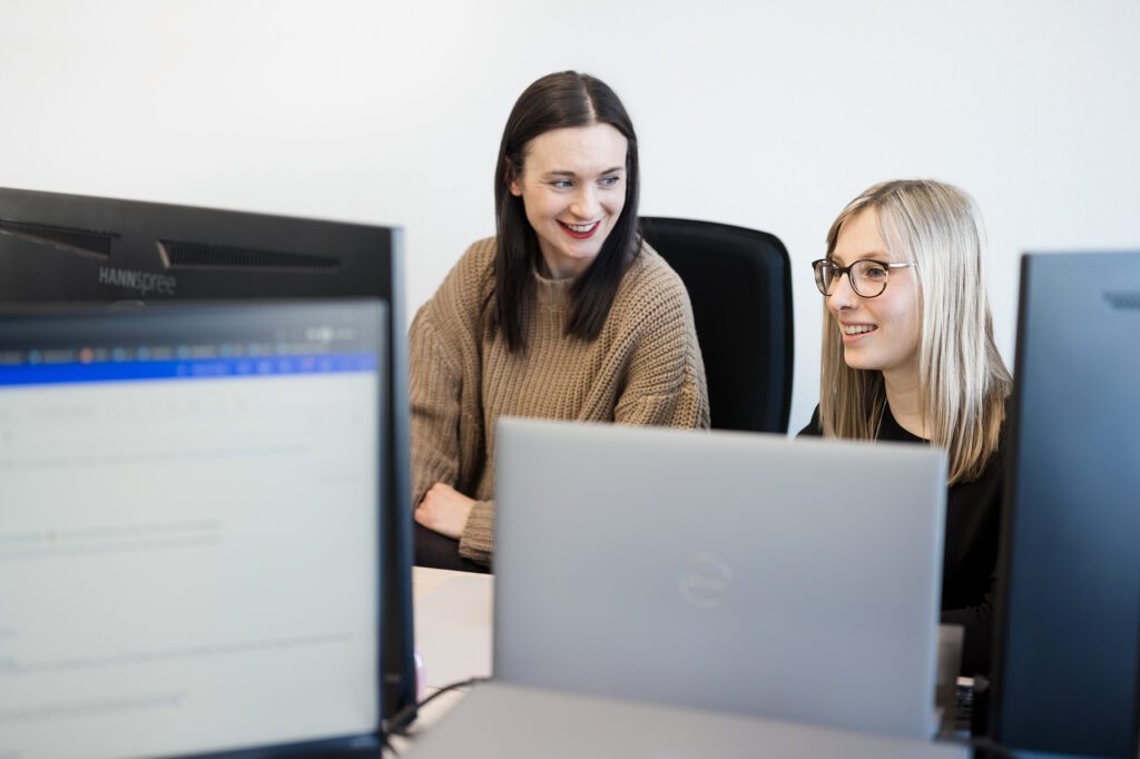 Zwei Frauen sitzen gemeinsam vor mehreren Monitoren im Büro, beide lächeln und wirken im Gespräch vertieft.