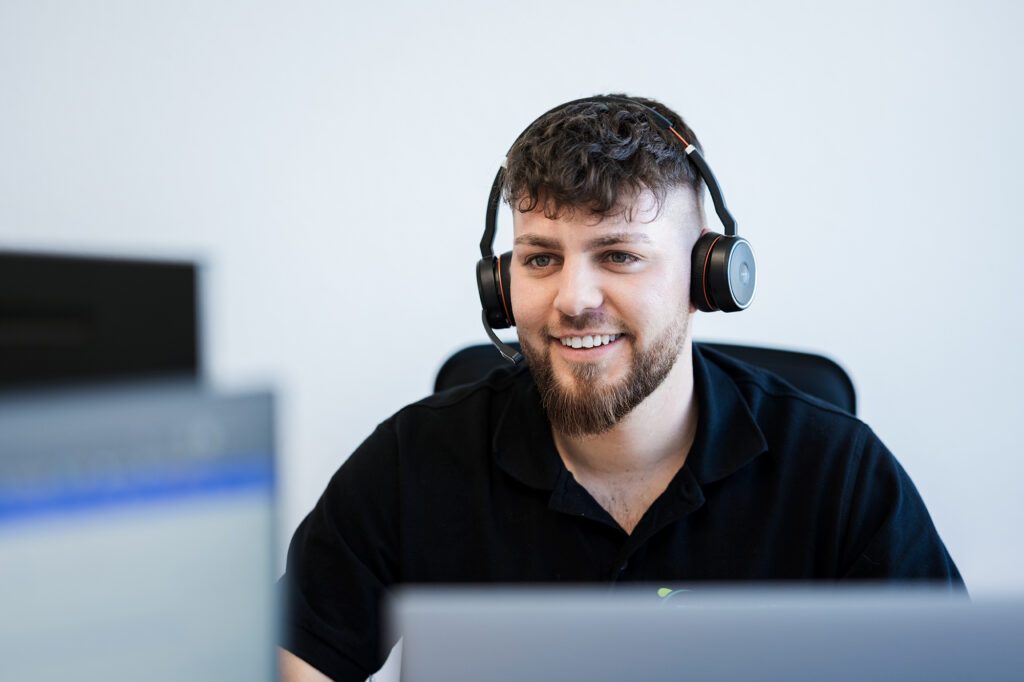 Kundenberater in schwarzem Poloshirt arbeitet mit Headset im Büro und blickt freundlich auf den Bildschirm.