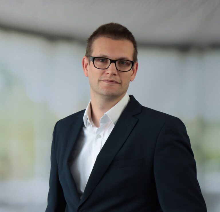 Porträt von André Lübke mit Brille im dunklen Anzug und weißem Hemd vor neutralem Hintergrund.