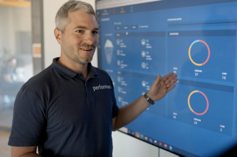 Ein Mitarbeiter mit grauen Haaren zeigt auf ein Dashboard mit Diagrammen auf einem großen Bildschirm und erklärt Inhalte.