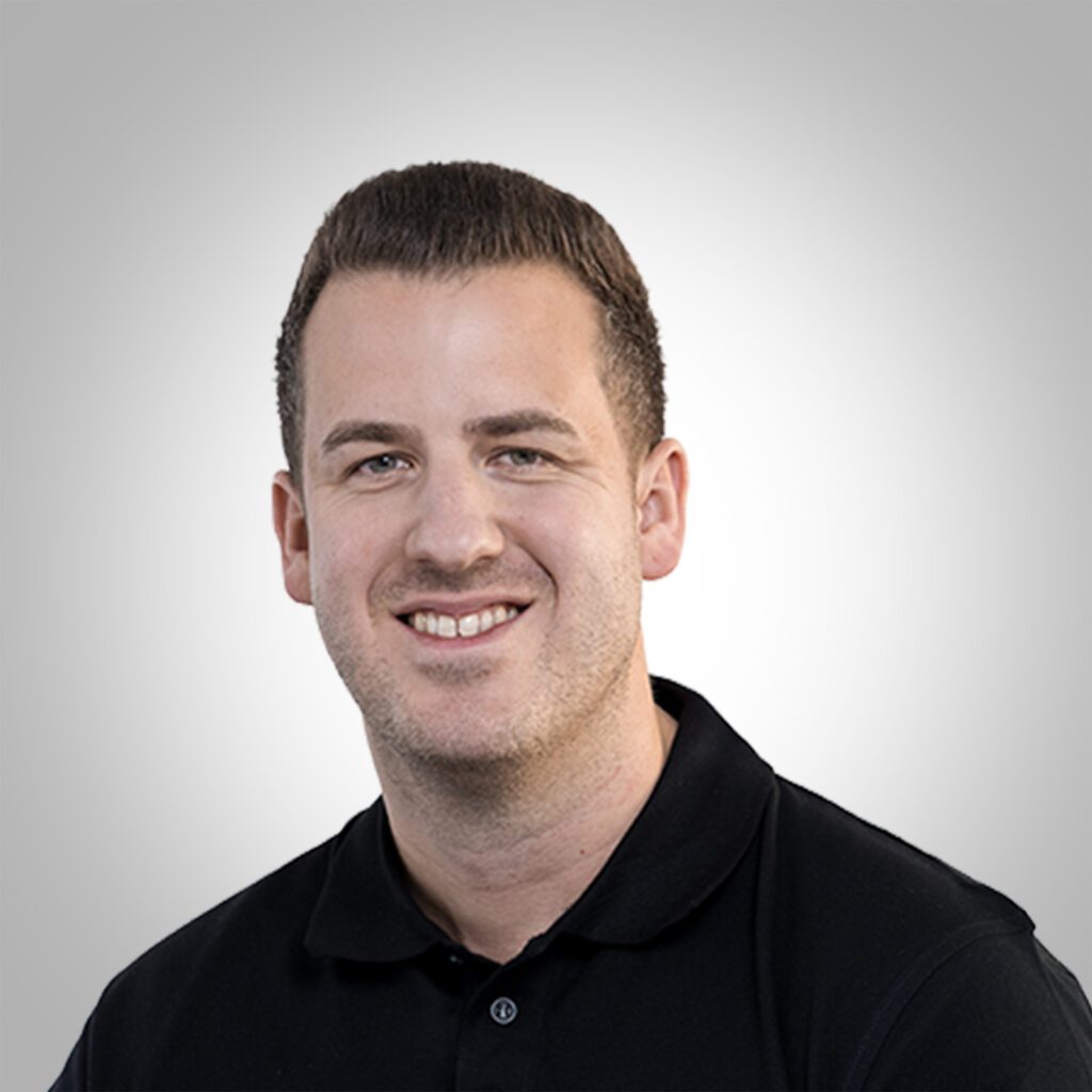 Porträt von Sascha Tiefenbach, Geschäftsstellenleiter, in in schwarzem Polohemd, lächelnd vor grauem Hintergrund.