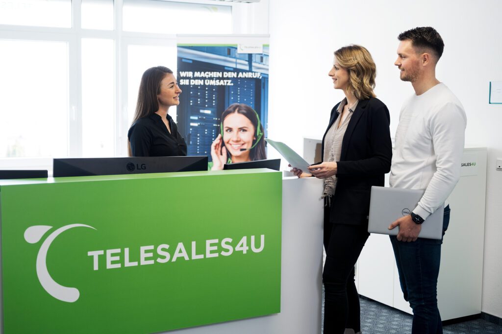 Empfangsszene in einem modernen Büro: Eine Frau hinter einem grünen Tresen mit Telesales4u-Logo spricht mit einem Mann und einer Frau mit Unterlagen in der Hand.